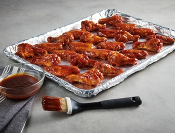 chicken wings lying on an aluminum foil lined baking sheet alongside a bowl of sauce and a basting brush