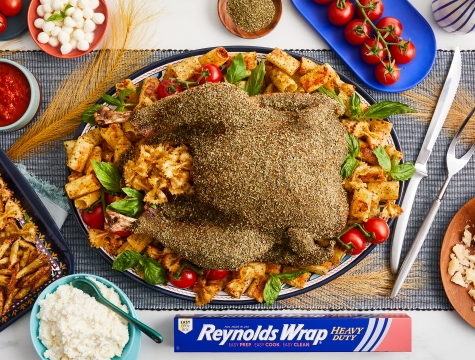 A cooked turkey on a plate, surrounded by pasta chips and tomatoes. A box of Reynolds Wrap heavy duty foil is nearby.