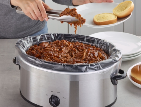 Person using tongs to scoop BBQ pulled pork from a lined slow cooker onto a bun
