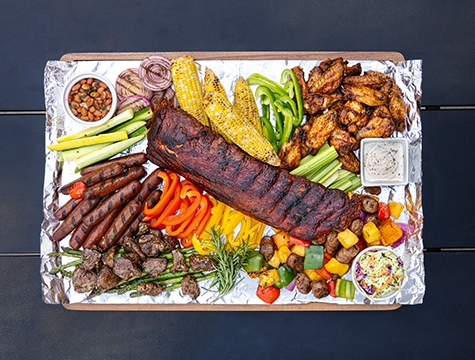 Rack of ribs, sausages, grilled corn on the cob and roasted vegetables sitting on foil lined wood cutting board