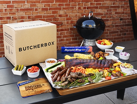 Butcher box package lying on an outdoor table surrounded by Reynolds Wrap Heavy Duty foil, meats and recipe cards