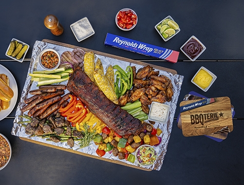 BBQ ribs, sausage, corn on the cob and roasted vegetables lying on an aluminum foil lined wood board