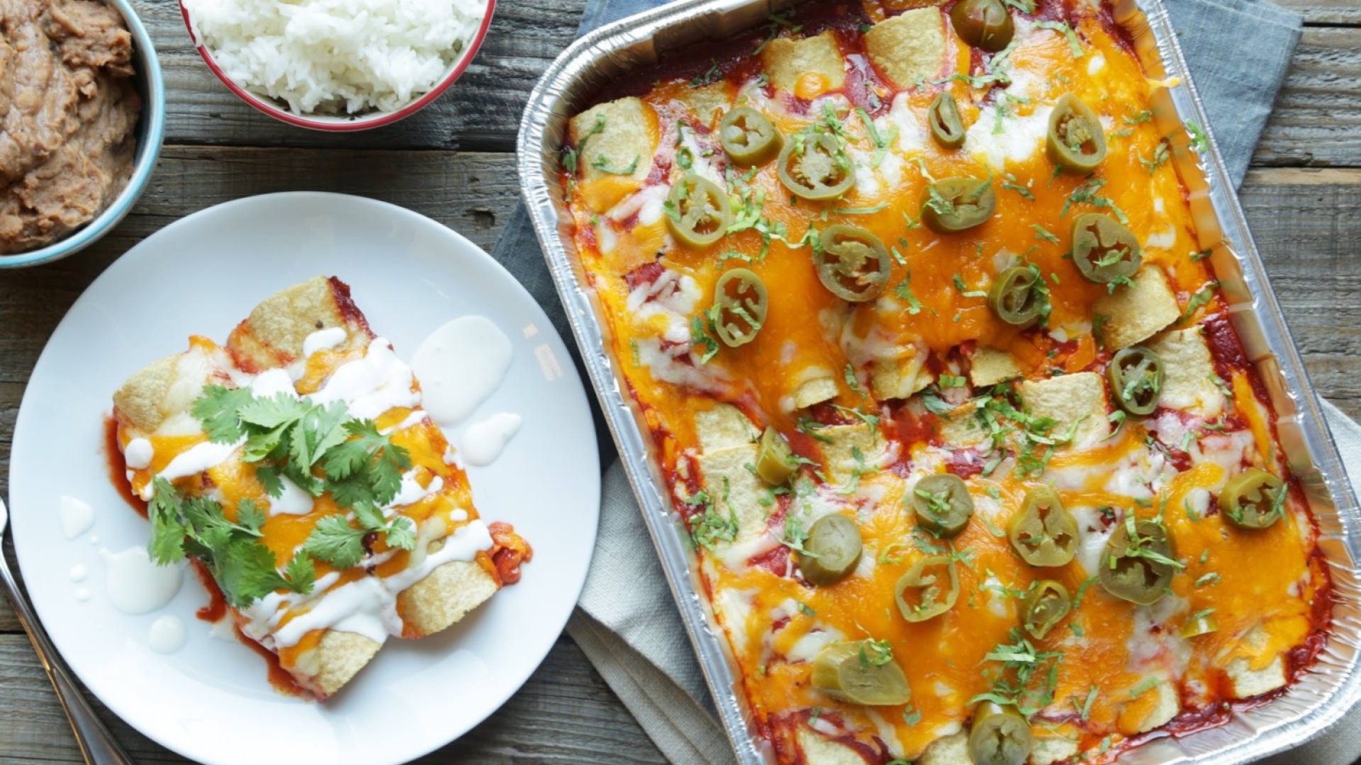 Enchiladas in a disposable aluminum foil bakeware pan on a table alongside a plate