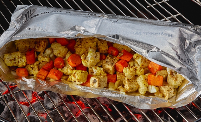 Roasted cauliflower and carrots in an open aluminum foil grill bag sitting on top of a hot grill