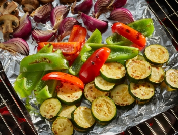A sheet of aluminum foil covering grill grates and topped with an assortment of sliced vegetables