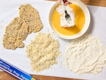 chicken cutlets sitting on a sheet of Reynolds Kitchens Countertop Prep Paper alongside a breading station of egg wash, flour and bread crumbs